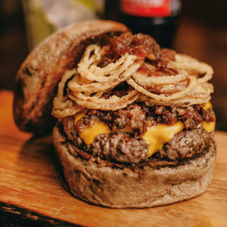 a large burger with the top bun leaning at its side to reveal a patty topped with cheese, deep fried onion rings, and ragu sauce.